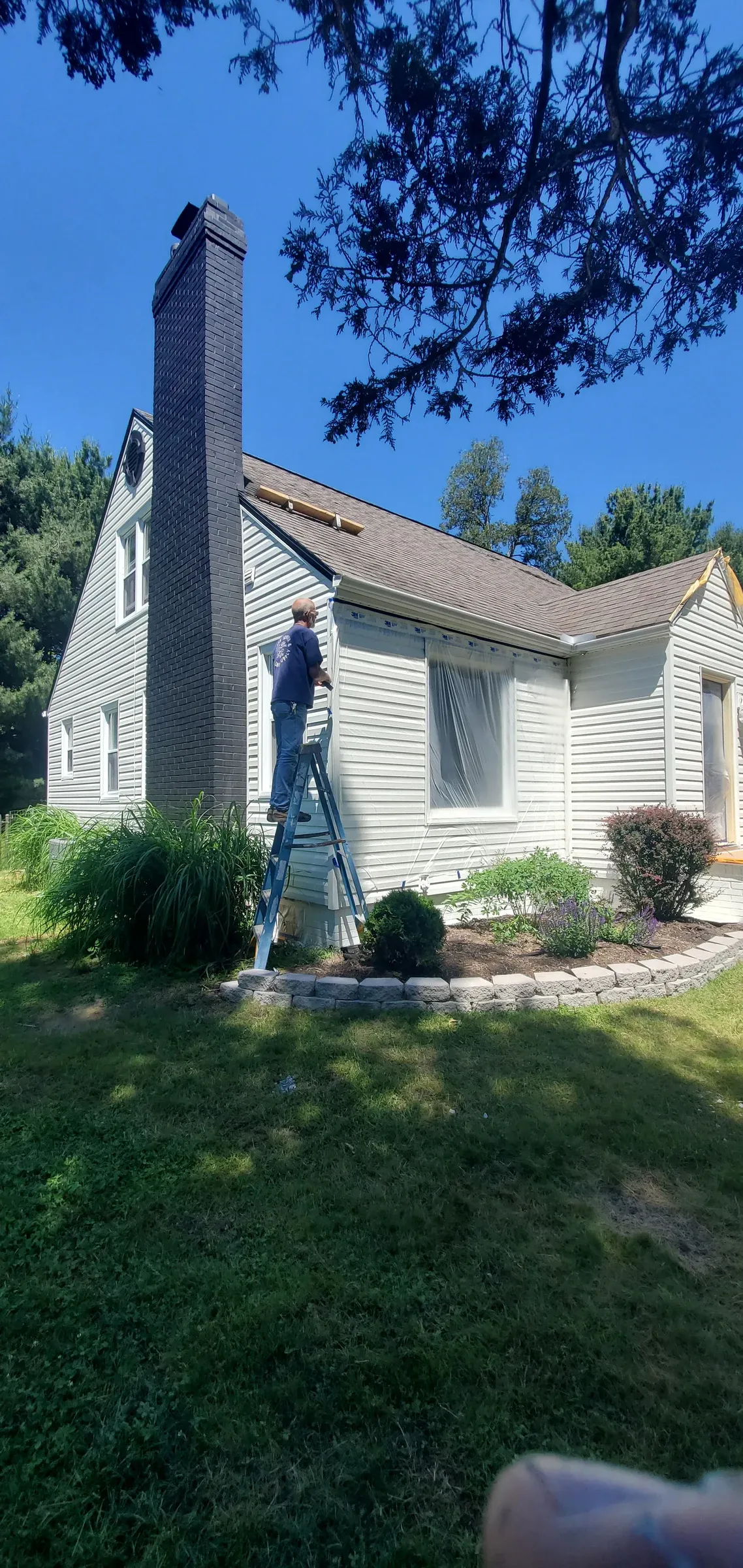 Painter on a ladder working along white siding beside a dark brick chimney